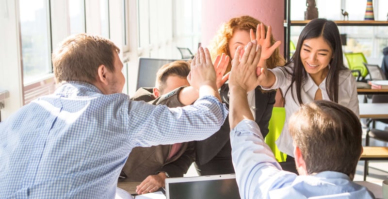 Teamwork: Ein Handschlag zwischen 5 Kolleg*innen über einem Schreibtisch mit Bildschirm und Notizblock. 